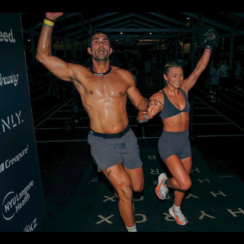 Two HYROX athletes holding hands while leaping forward during a race, with sponsor logos like HYROX, MANLY, and NYU Langone Health visible in the background.